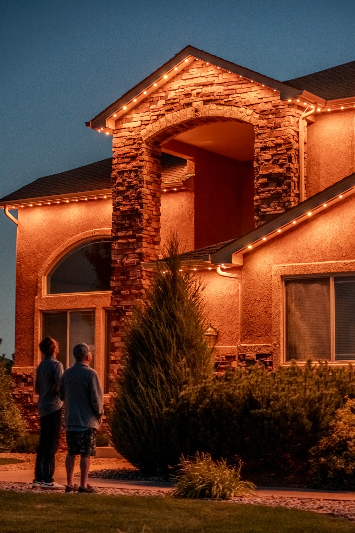 Warm white roofline lighting on a stone-front home with homeowners outside